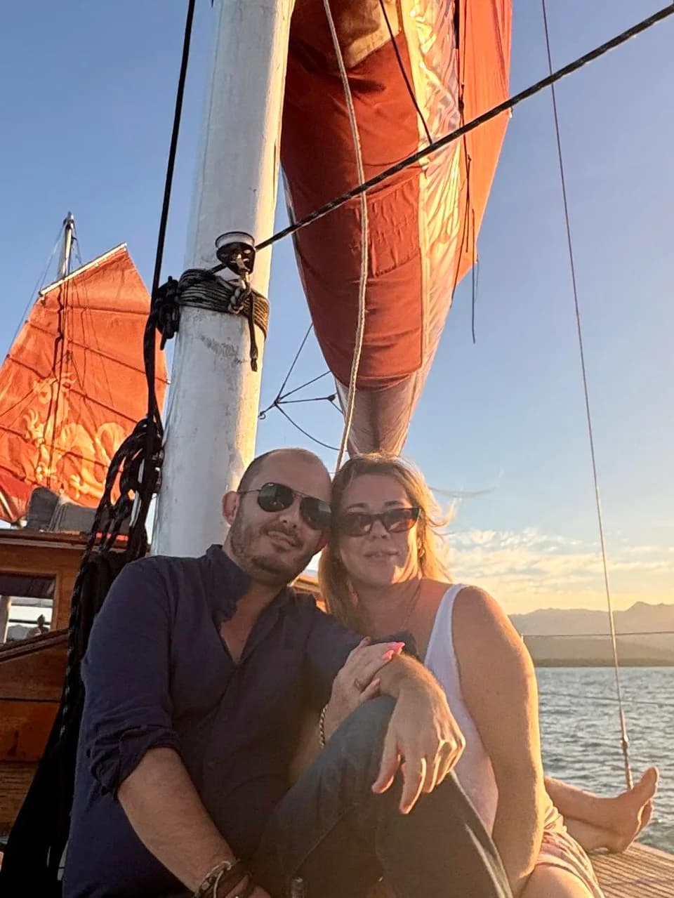 Two people aboard a sailing boat beneath orange sails at sunset.